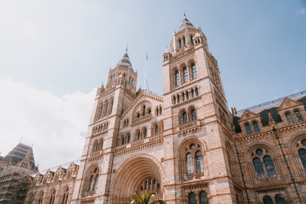 A grand historic building with two tall clock towers, arched windows, and detailed stonework, set against a partly cloudy sky.