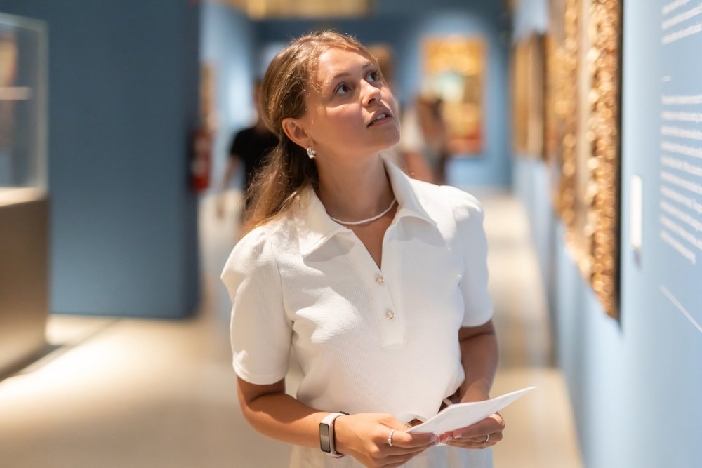 A woman in a white shirt holds a paper and looks thoughtfully at artwork in a museum with blue walls and framed displays.