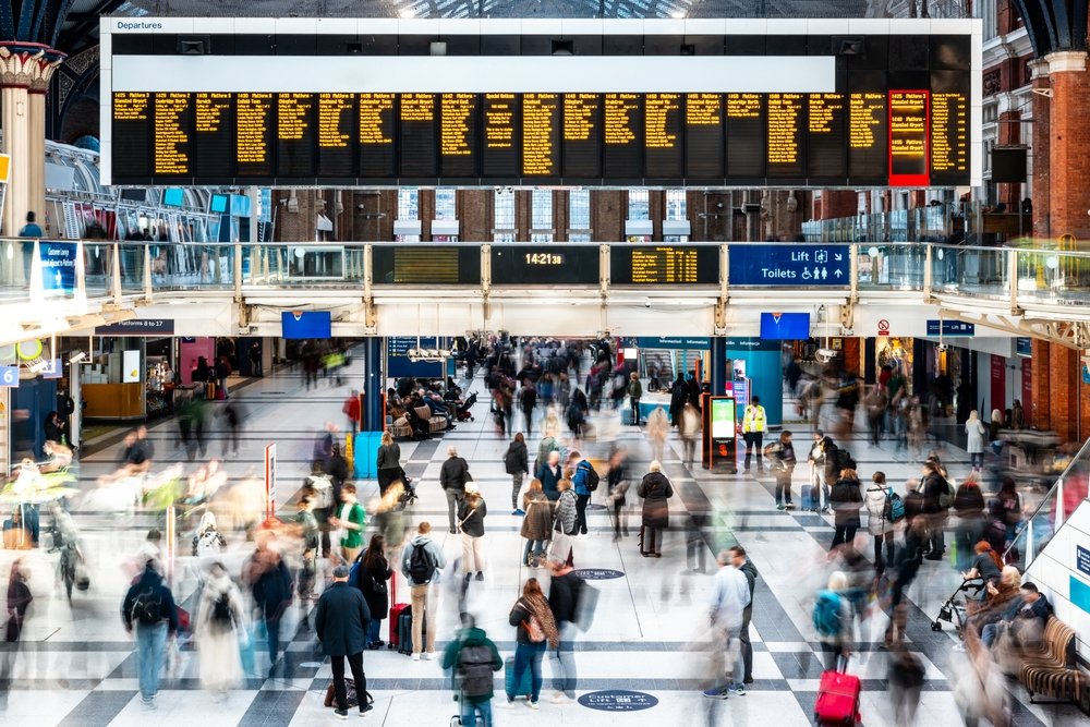 A busy train station with blurred crowds of people walking and standing beneath a large digital departure board displaying train schedules. Signs for exits and toilets are visible above the crowd.