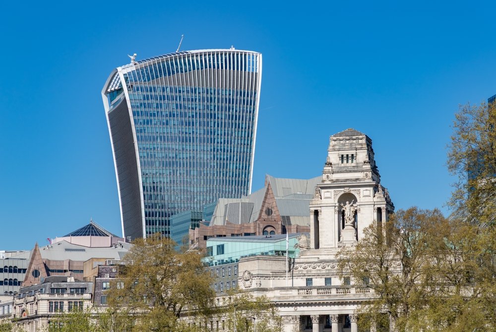 A modern, curved skyscraper known as the Walkie Talkie building rises behind historic stone buildings and leafy trees in London, under a clear blue sky.