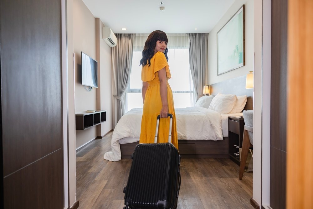 A woman in a yellow dress stands in a modern hotel room, looking back while pulling a black suitcase. The room features a large bed, wall-mounted TV, wooden floor, and sunlight streaming through curtains.