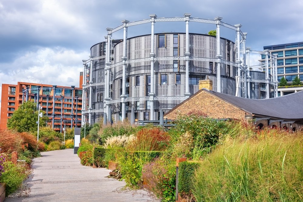 A modern circular building with a metallic frame and glass facade stands next to a brick structure, surrounded by lush greenery and a paved pathway under a cloudy sky.