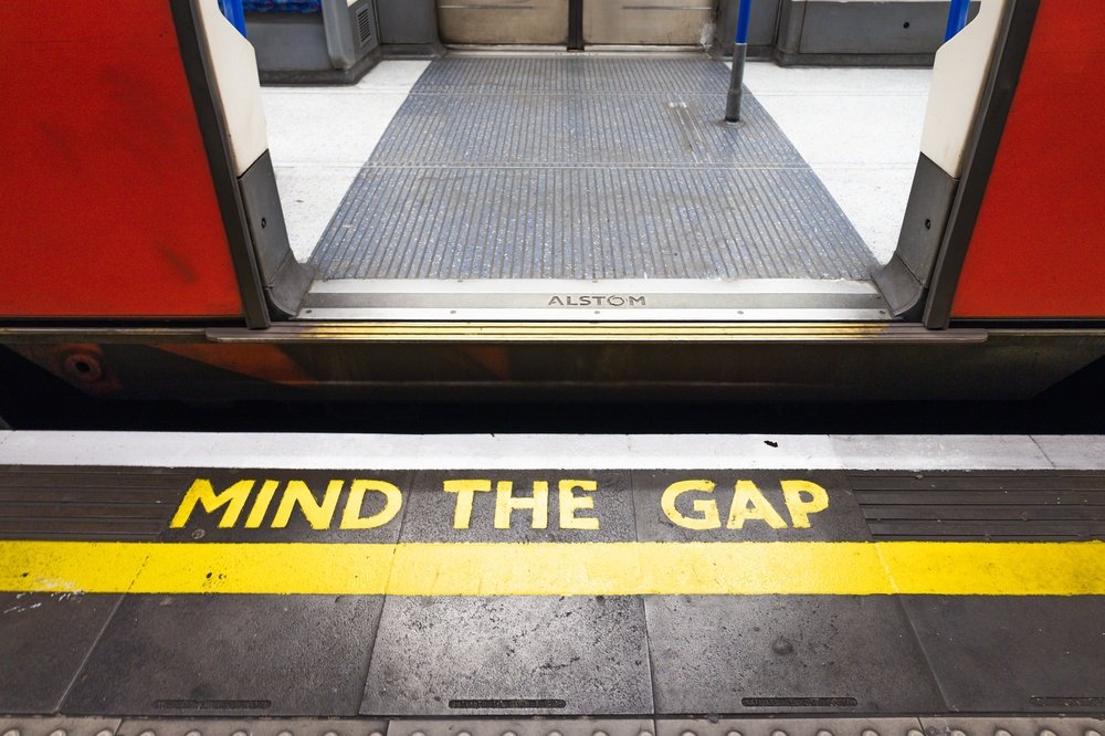 Open train doors at a subway platform with the words "MIND THE GAP" painted in yellow on the ground near the edge, highlighting the gap between the train and the platform.
