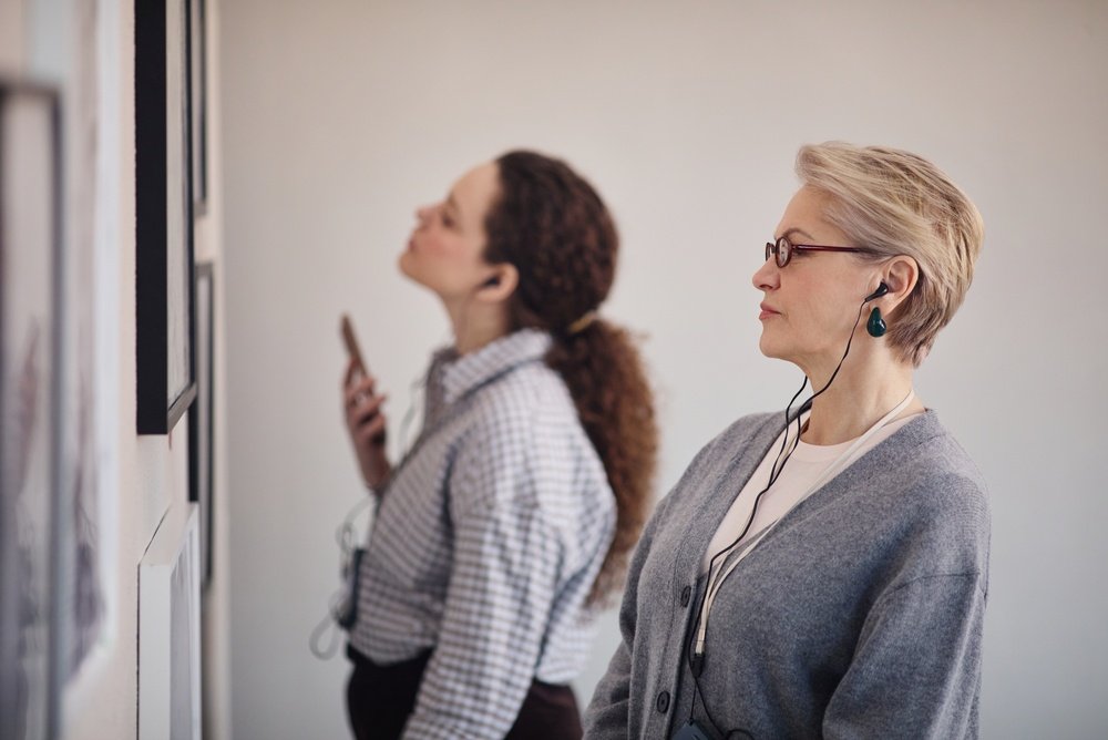 Two women wearing headphones stand in an art gallery, closely observing framed artworks on the wall. One holds a smartphone, and both appear focused and engaged with the exhibits.