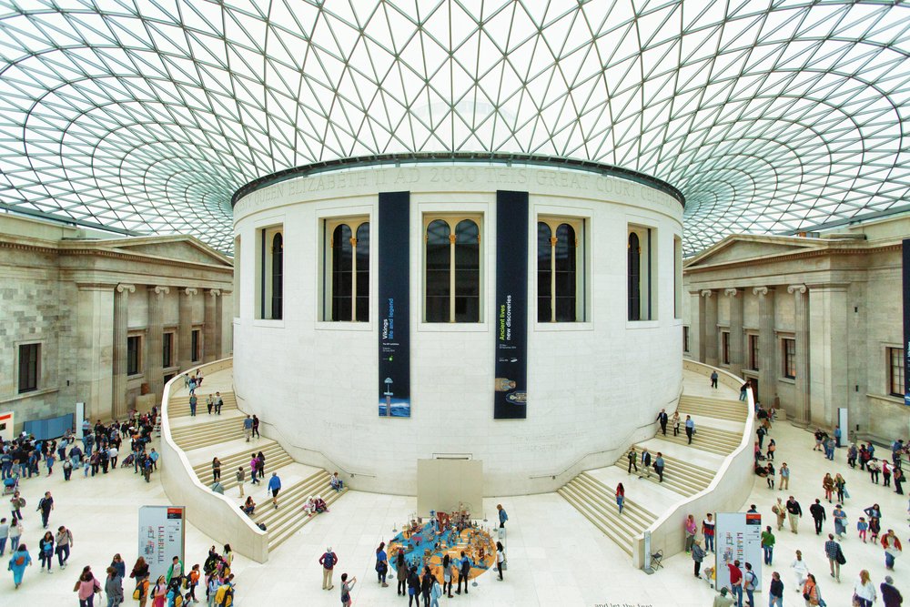 A large, bright atrium with a glass roof and white walls in the British Museum; people walk around and gather near exhibits and grand staircases on both sides.