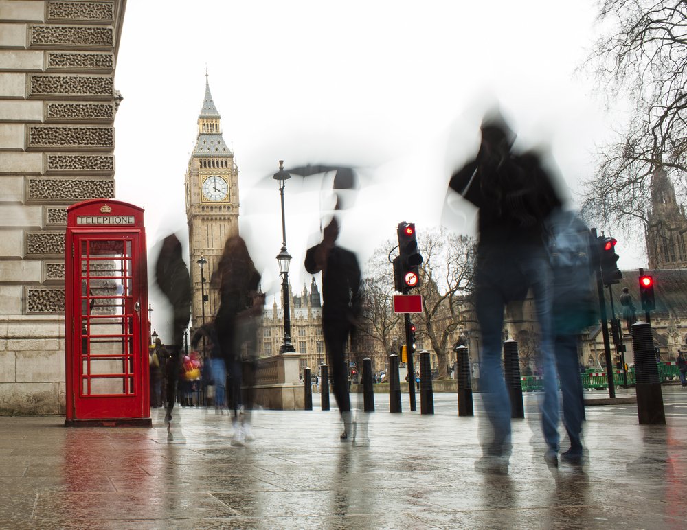 Blurred pedestrians walk past a classic red phone booth near Big Ben in London on a wet, overcast day, with traffic lights and historic buildings visible in the background.