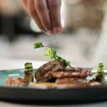 A chef’s hand sprinkles garnish over a plated gourmet dish—grilled meat, vegetables, and decorative greens on a blue-green plate—capturing the essence of the Best Places to Eat Near Covent Garden Before a Theatre Show.
