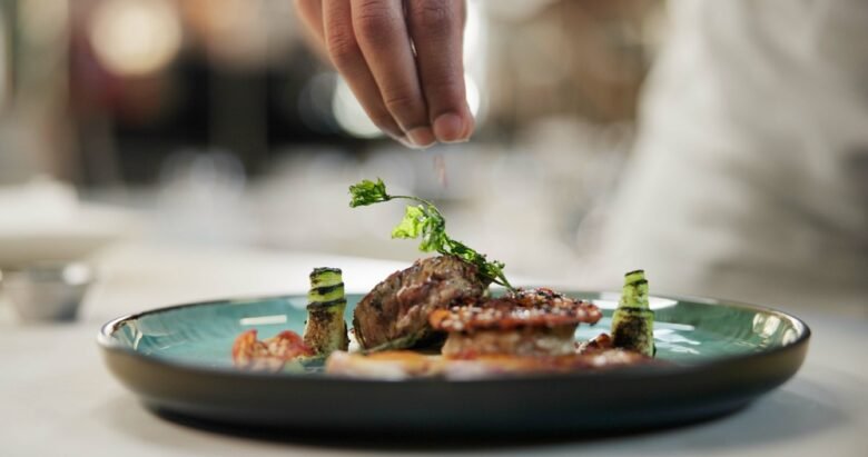 A chef’s hand sprinkles garnish over a plated gourmet dish—grilled meat, vegetables, and decorative greens on a blue-green plate—capturing the essence of the Best Places to Eat Near Covent Garden Before a Theatre Show.