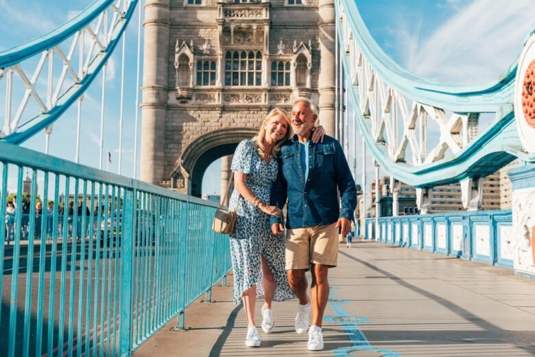 A smiling couple walks hand in hand on Tower Bridge in London on a sunny day. The woman wears a blue patterned dress, and the man wears a denim shirt and shorts. The iconic bridge structure is visible behind them.