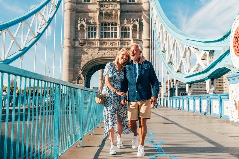 A smiling couple walks hand in hand on Tower Bridge in London on a sunny day. The woman wears a blue patterned dress, and the man wears a denim shirt and shorts. The iconic bridge structure is visible behind them.