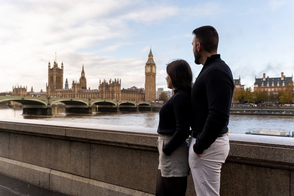 A man and woman stand by the River Thames, looking toward the Houses of Parliament and Big Ben in London on a cloudy day.