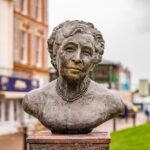 A stone bust of an elderly woman with short, wavy hair and a pearl necklace, displayed outdoors on a pedestal, with blurred buildings and greenery in the background.