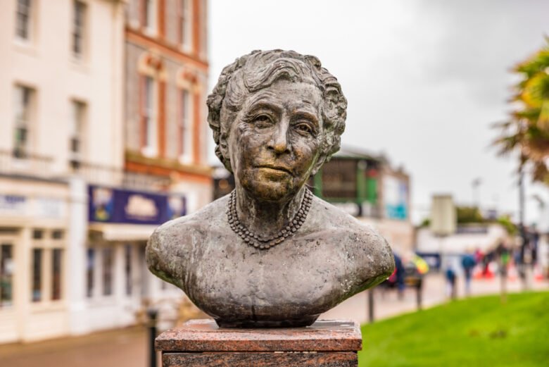 A stone bust of an elderly woman with short, wavy hair and a pearl necklace, displayed outdoors on a pedestal, with blurred buildings and greenery in the background.