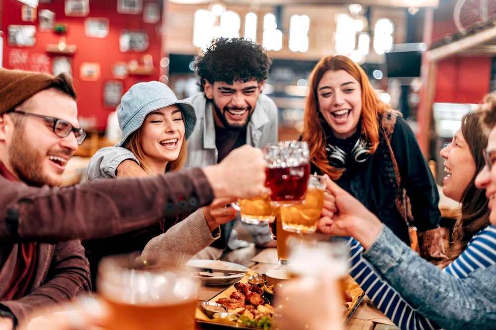 A group of young adults sit around a table in a lively restaurant or bar, laughing and toasting with drinks, surrounded by food and a warm, cheerful atmosphere.