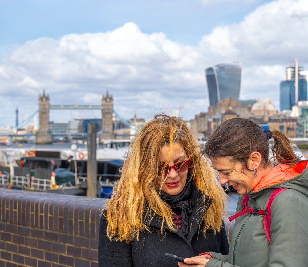 Two women stand by a brick wall overlooking the Thames in London, smiling and looking at a phone. The Tower Bridge and city skyscrapers are visible in the background under a partly cloudy sky.