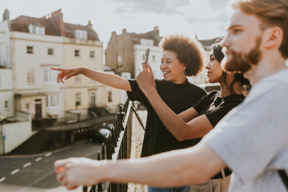 Three young adults stand outdoors near a railing, with one person pointing excitedly while another takes a photo with a phone. The group enjoys a sunny day in a residential area with houses in the background.
