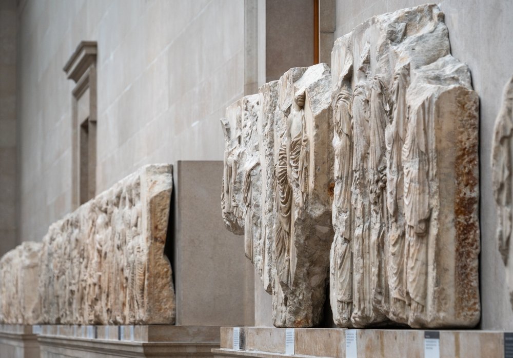 Marble relief sculptures depicting figures in draped clothing are displayed along a museum wall, with soft lighting highlighting the intricate details of the ancient carvings.