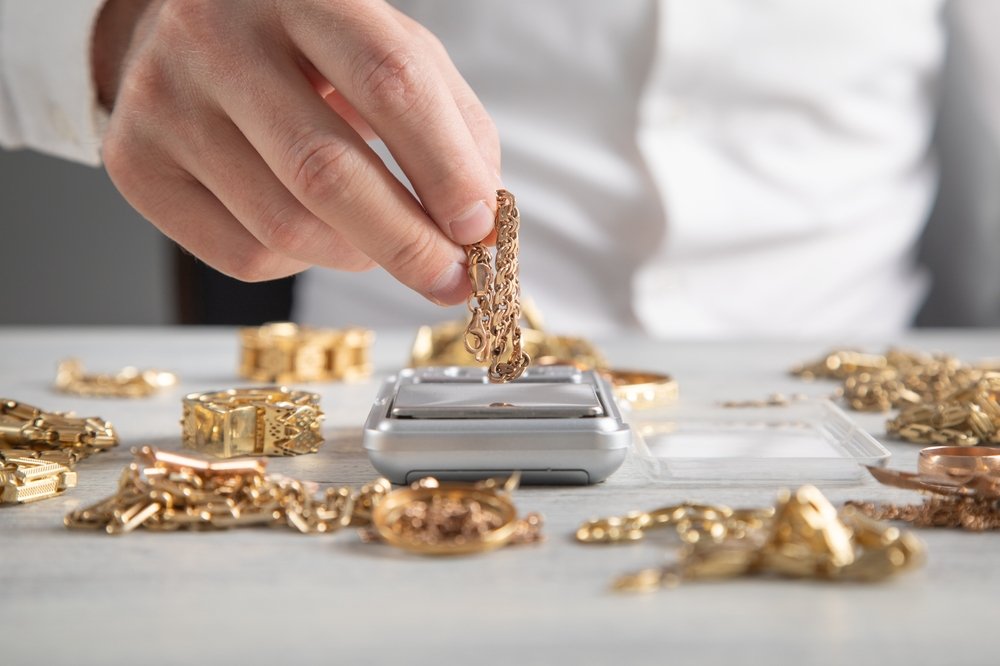 A person in a white shirt places a gold chain on a digital scale, surrounded by various gold jewelry pieces on a light-colored table.