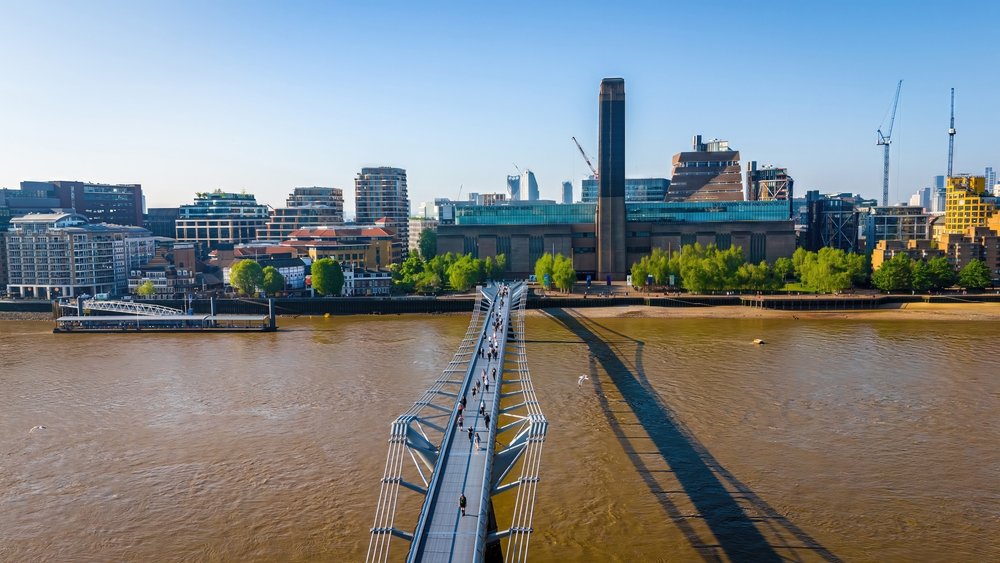 Aerial view of the Millennium Bridge spanning the River Thames in London, with people walking across. The Tate Modern art museum and modern city buildings are visible on the opposite bank under a clear blue sky.