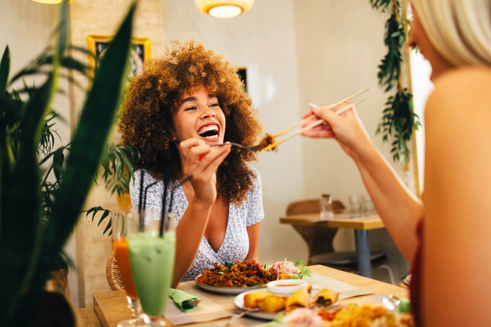 A woman with curly hair smiles and laughs while another person, partially visible, feeds her with chopsticks at a restaurant table filled with food and a green drink.