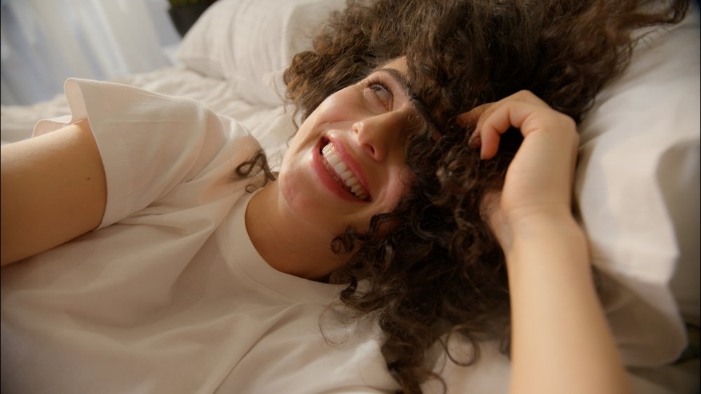 A woman with curly hair lies on a bed, smiling and looking upwards. She is wearing a white t-shirt, and natural light is softly illuminating her face.