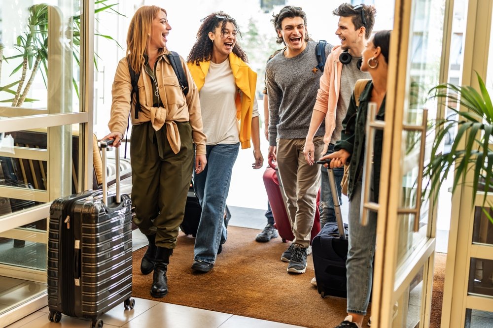 Five young adults with luggage walk through a doorway, smiling and laughing together, suggesting they are arriving at or leaving a hotel or apartment. Green plants are visible near the entrance.