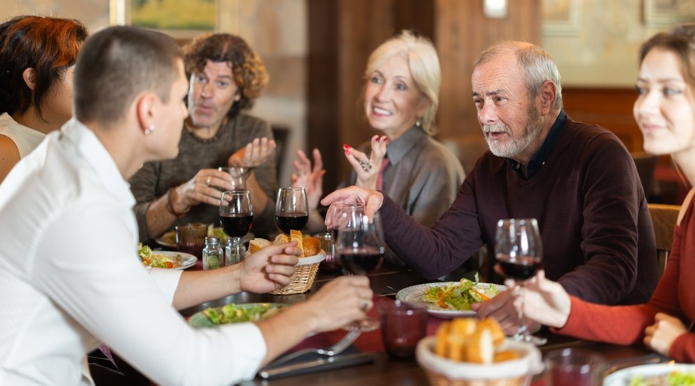 A group of six adults sit around a dining table, enjoying wine and conversation. Plates of salad and baskets of bread are on the table, and everyone appears engaged and cheerful.