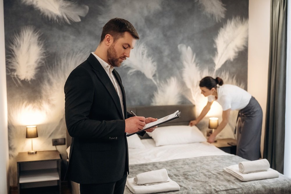 A man in a suit writes on a clipboard while a woman in uniform makes a neatly arranged bed in a hotel room with a feather-patterned accent wall.