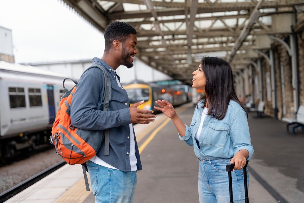 A man with a backpack and a woman with a rolling suitcase stand on a train platform, smiling and talking to each other as trains are visible in the background.