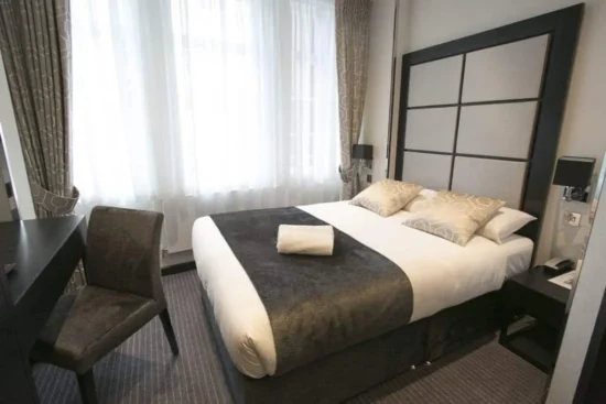 A modern hotel room near Liverpool Street Station with a double bed, two pillows, and a folded towel. The room features a desk with chair, bedside tables, and large windows with sheer curtains letting in natural light.