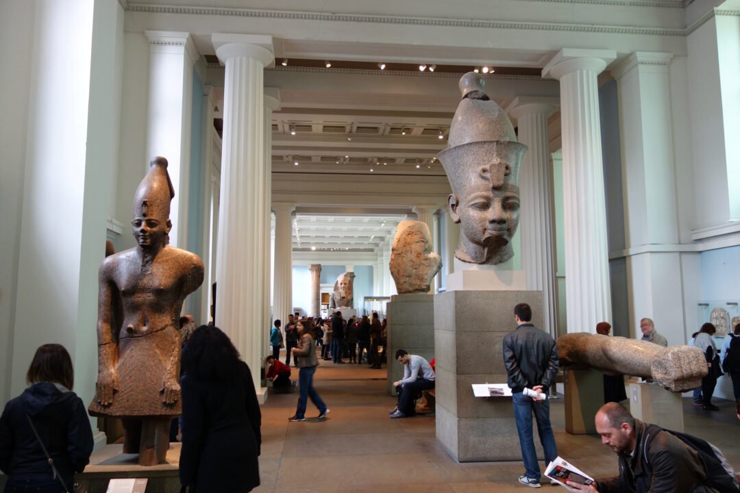 A museum hall with tall white columns displays large ancient Egyptian statues and sculptures; visitors, some staying at hotels near British Museum, are walking, reading signs, and sitting throughout the space.