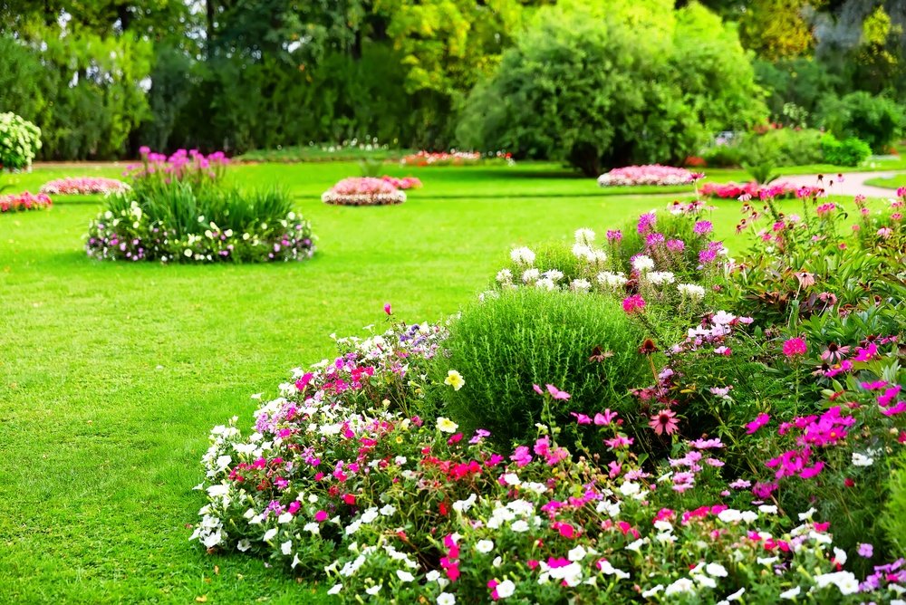 A vibrant garden with neatly trimmed grass, colorful flower beds, green shrubs, and blooming plants in various shapes, surrounded by lush trees and greenery in the background.