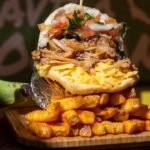 A sandwich stuffed with shredded meat, grated cheese, diced tomatoes, onions, and herbs, sits atop a pile of golden, seasoned potato fries on a wooden tray.