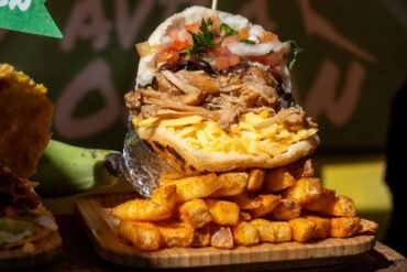 A sandwich stuffed with shredded meat, grated cheese, diced tomatoes, onions, and herbs, sits atop a pile of golden, seasoned potato fries on a wooden tray.