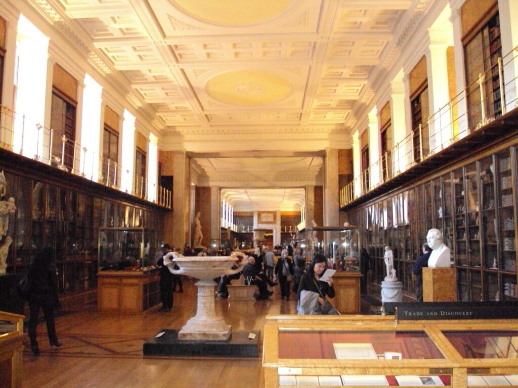 A grand museum hall with high ceilings, glass display cases, sculptures, and visitors walking around and viewing exhibits in a well-lit, elegant space.