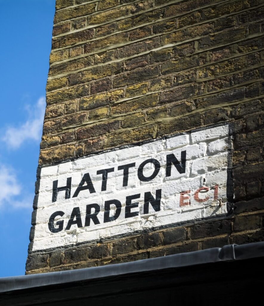 A white street sign reading "HATTON GARDEN EC1" is painted on a brick wall. The sign is slightly worn, and part of a roof edge is visible below. The sky is blue with a few clouds.