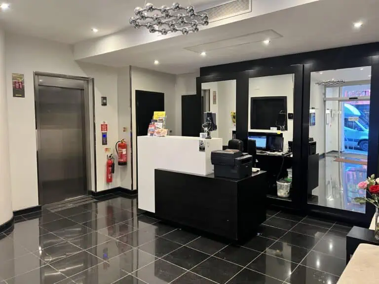 Modern hotel reception area with a black and white color scheme, glossy black tile floor, sleek reception desk, mirrors, computer, elevator, and fire safety equipment—ideal for travelers seeking hotels near Liverpool Street Station.