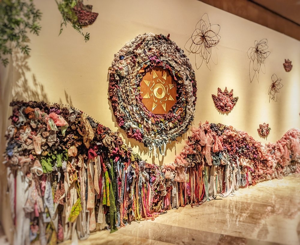 Textile art installation on a wall featuring a large circular sun motif made of colorful fabric scraps, with waves and flower shapes, and strips of fabric hanging below—an inviting sight for anyone inspired by the Bloomsbury Guide for First-Time Visitors.