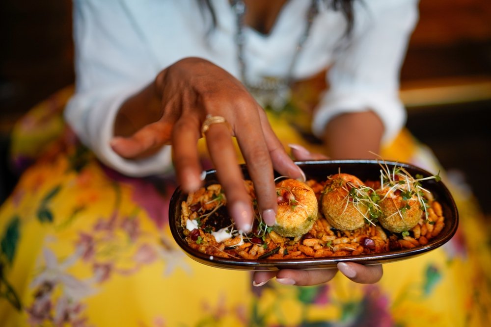A person wearing a white top and yellow floral skirt holds a plate of three round, garnished appetizers on a bed of crispy grains, while reaching to pick one up.