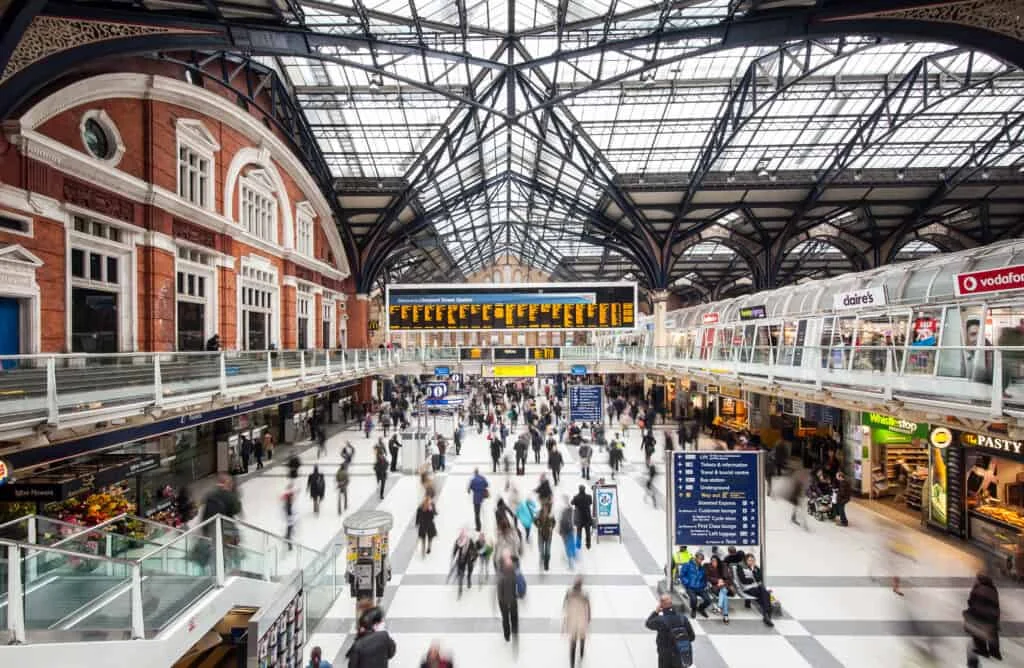Busy train station interior with a glass roof, people walking in various directions, shops along the sides, and large digital departure boards displaying train times above the crowd.