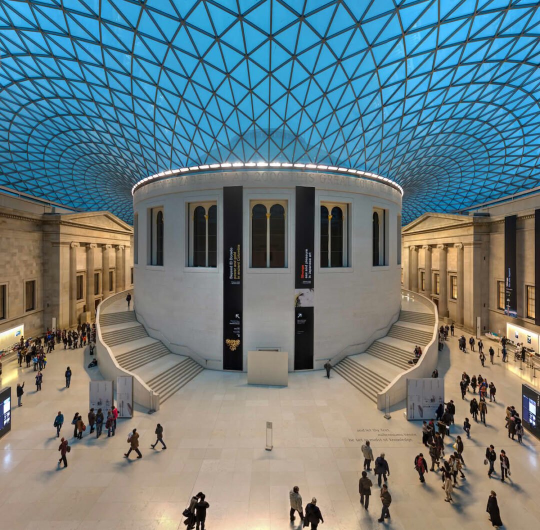 A spacious museum interior with a large circular stone building in the center, surrounded by wide stairs. The glass-paneled roof forms a blue geometric pattern above, just steps from hotels near British Museum where visitors can stay.