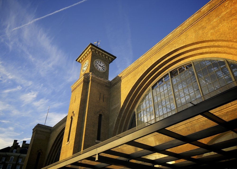A yellow-brick building with a large arched window and a clock tower, pictured from below. The sky is blue with wispy clouds and a contrail, and sunlight highlights the building’s façade.