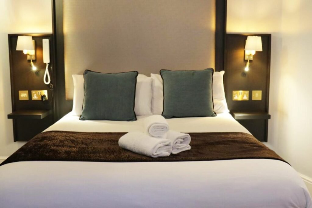 A neatly made hotel bed with two green cushions, brown throw, and two rolled white towels on top. The headboard is flanked by two bedside lamps and built-in nightstands with phones and power outlets.