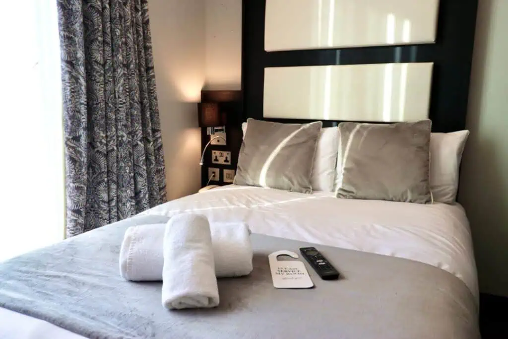 A neatly made bed with gray and white bedding, two folded towels, a TV remote, and a door hanger on top. Curtains and a small lamp are visible beside the bed.