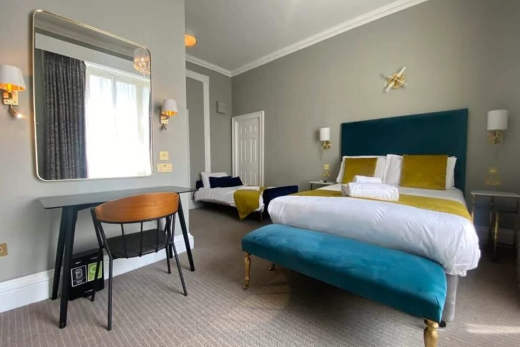 A modern hotel room with a double bed, teal headboard, yellow pillows, a teal bench, a desk with a chair, a large mirror, and a single bed by the window. The room is bright with neutral walls and carpet.