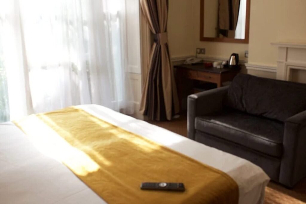 A sunlit hotel room with a bed covered by a yellow runner, a remote control on the bed, a gray sofa, a desk with a kettle, a mirror, and large windows with sheer curtains.