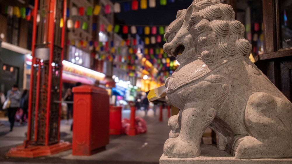 A stone lion statue sits in the foreground of a lively street at night, decorated with colorful lanterns hanging above, with blurred people and buildings in the background.