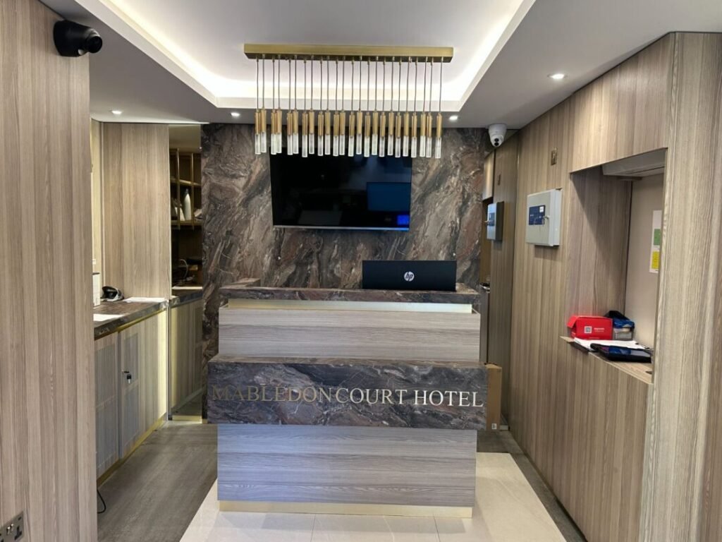 Modern hotel reception area with wood paneling, marble decor, a hanging light fixture, a TV screen, and a front desk with a computer. The words “Mabledon Court Hotel” are on the desk, marking the welcoming result of Crown Group’s Mabledon Court Hotel Refurbishment Complete.