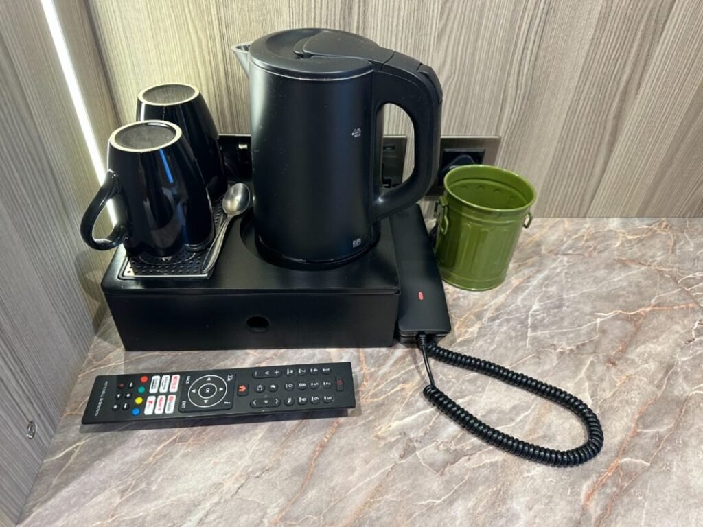 A black electric kettle, two black mugs, a green bucket, and a TV remote sit on a marble countertop with a telephone handset, against a wood-paneled wall—a cozy scene from Crown Group’s Mabledon Court Hotel Refurbishment Complete.