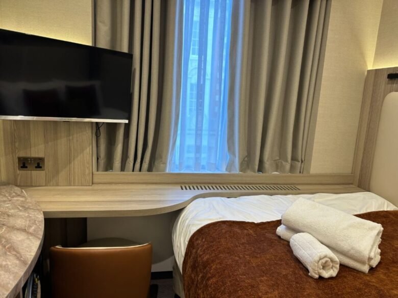 A neatly made hotel room with a bed, brown blanket, and white towels on top, a desk with a chair, a wall-mounted TV, and a window with beige curtains letting in daylight showcases the comfort and modern touches following Crown Group’s Mabledon Court Hotel Refurbishment Complete.